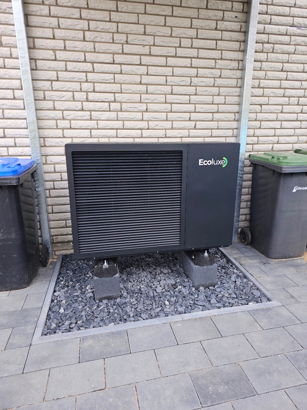 Eine schwarze Ecolux Wärmepumpe steht auf Steinen zwischen zwei Mülltonnen vor einer hellen Backsteinwand