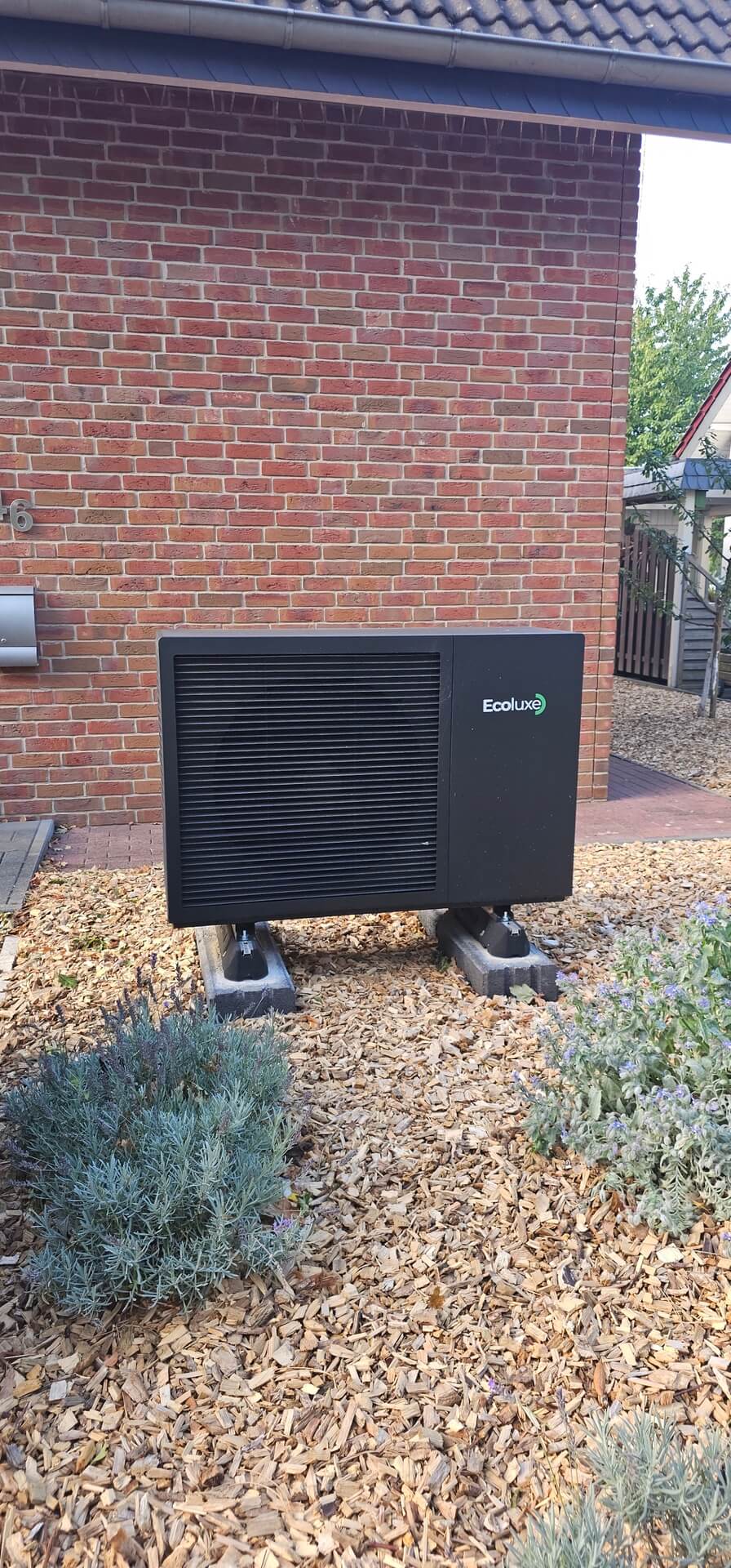 Eine schwarze EcoLux Wärmepumpe steht vor einer roten Ziegelhauswand im Garten