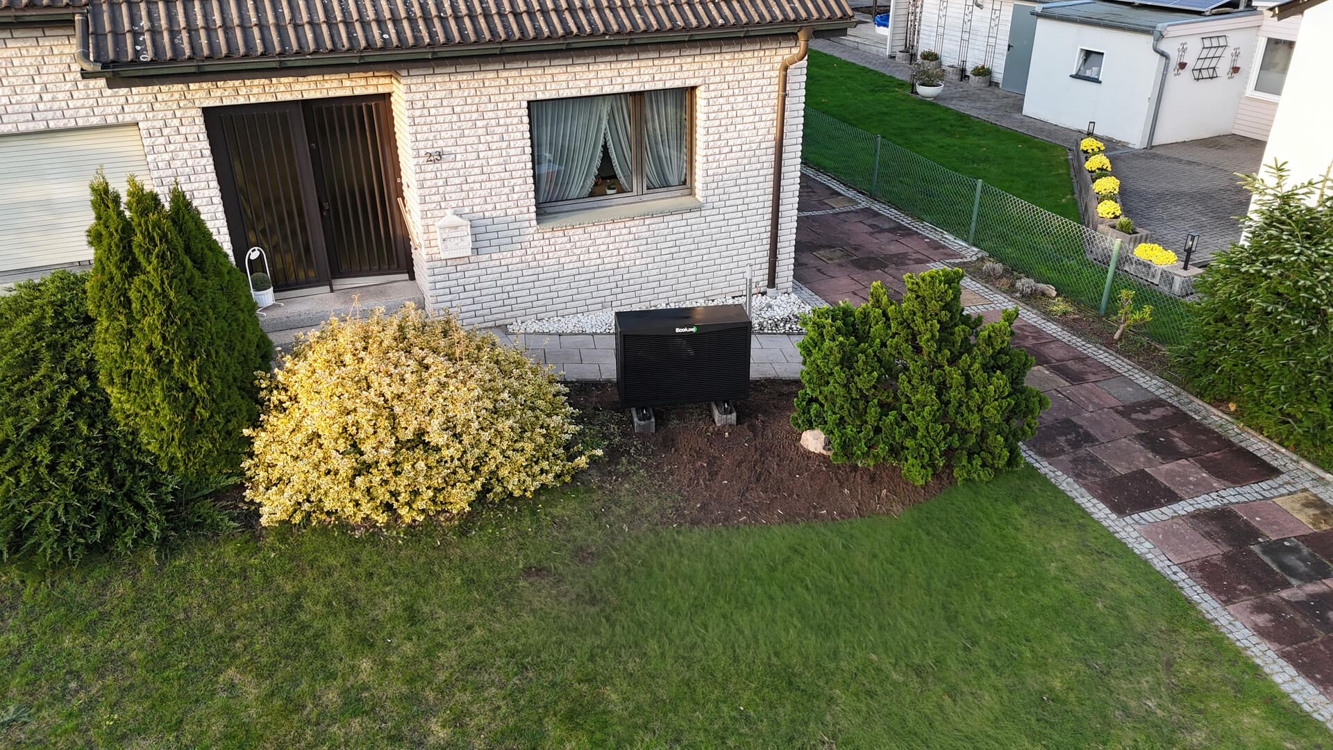 Vor einem weißen Haus steht ein schwarzes Solarpanel im Garten zwischen grünen Büschen